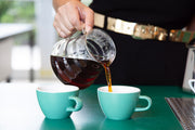 Coffee is being poured into one of two small teal ceramic cups on a teal counter.